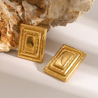 Gold rectangular earrings on a textured surface with a rock background