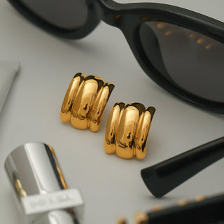 Gold hoop earrings with sunglasses and a silver lip gloss on a light gray background
