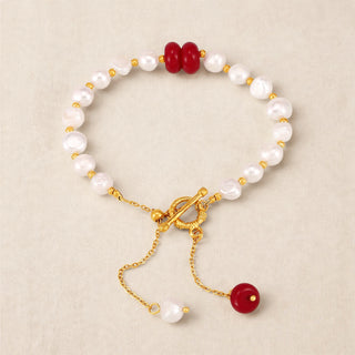 A gold-tone beaded bracelet with white freshwater pearls, accented by two red beads at the top and a dangling red bead charm at the bottom, displayed on a neutral beige background.