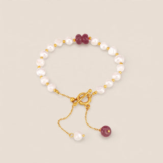 A delicate beaded bracelet with white baroque pearls, gold-toned metal links, a toggle clasp, and two small dark red gemstone beads, shown on a neutral beige background.