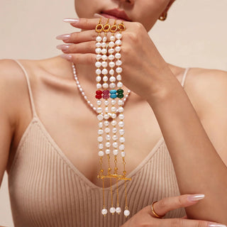 A close-up of a model wearing a multi-strand pearl necklace with gold-toned chains, colorful bead accents, and dangling charms, styled with a beige ribbed tank top.