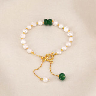 A gold-tone beaded bracelet with white freshwater pearls, featuring a double green stone bead, a dangling single green bead, and a gold toggle clasp, displayed on a neutral beige background.